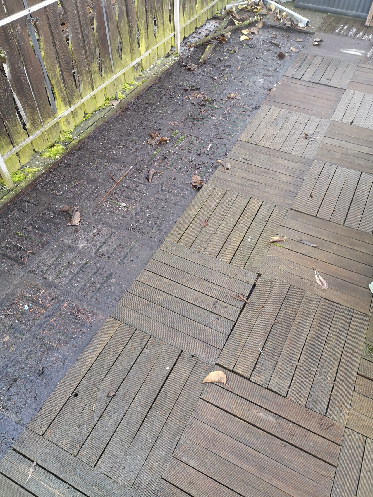 Wooden deck with rectangular boards; next to it a dirty soil strip with leaves and moss by the fence.