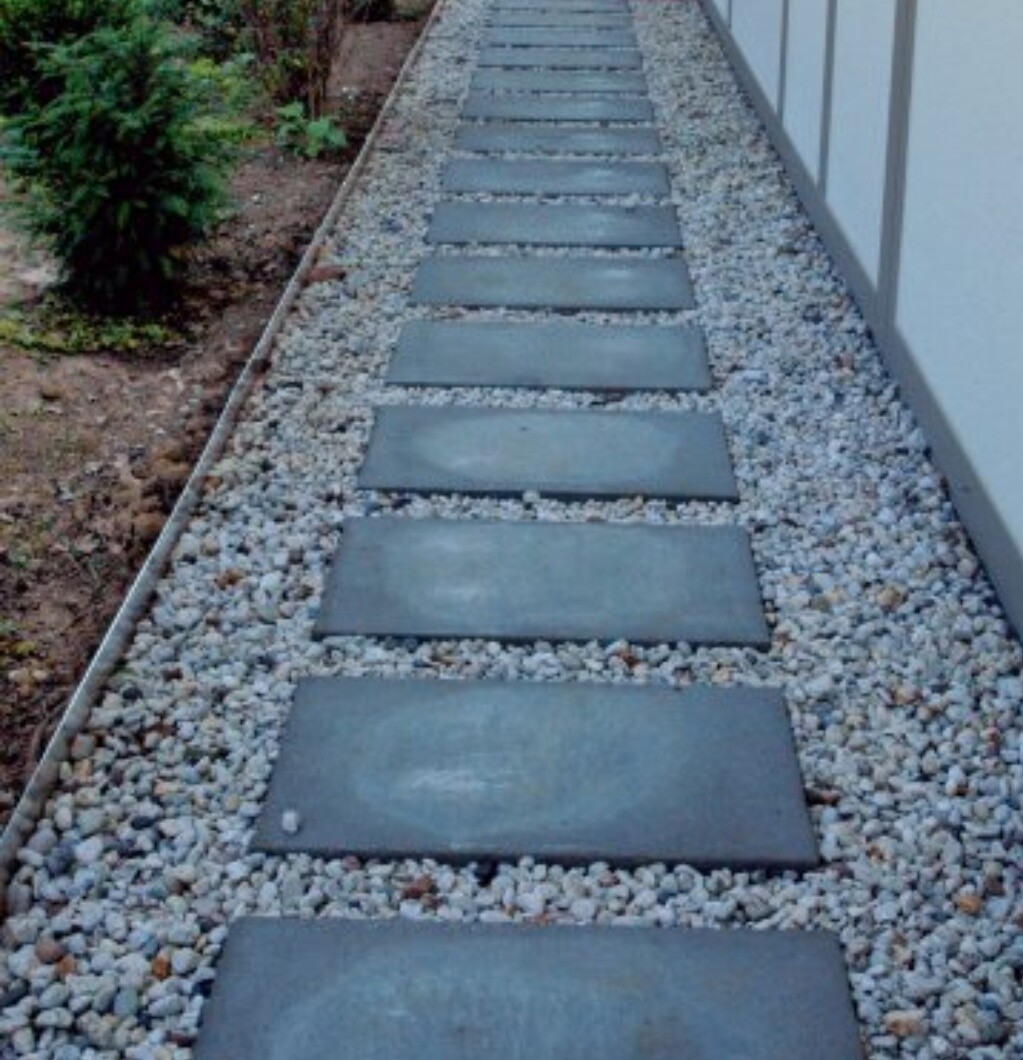 Garden path made of square stone slabs on gravel, next to a house wall, shrubs on the left.