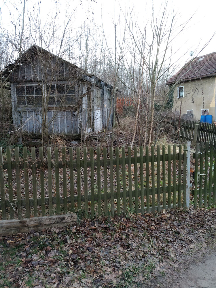 Abandoned wooden hut behind a wooden fence, surrounded by bare trees and autumn leaves.