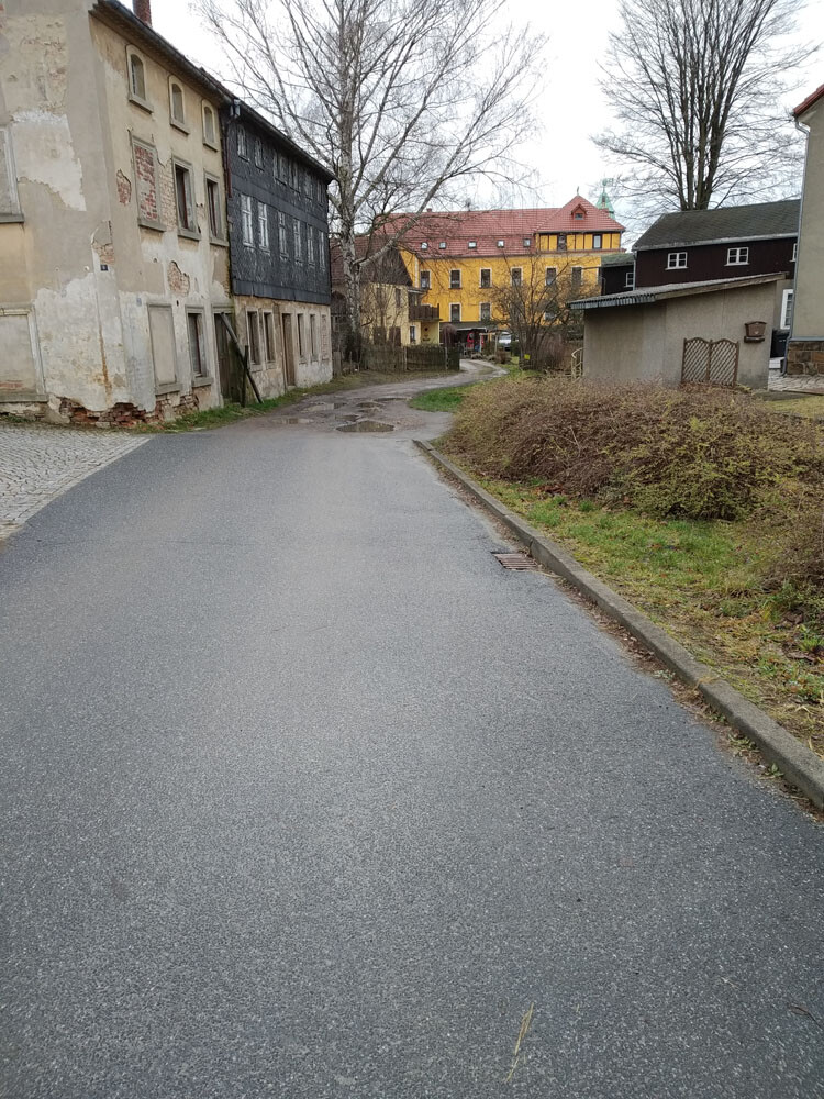 Small village street with old houses on the left, wet asphalt, yellow building in the distance.