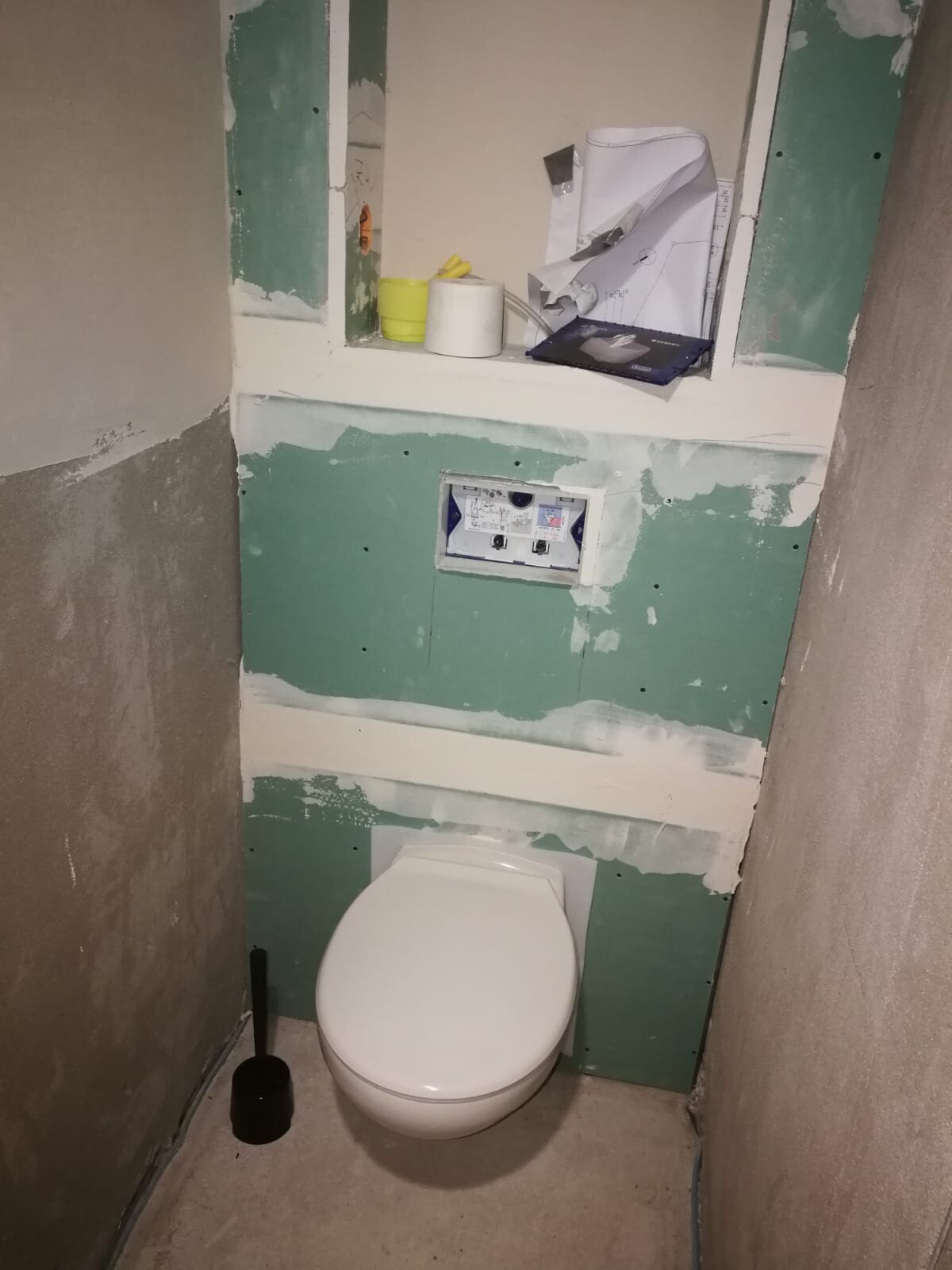 Unfinished bathroom with green drywall, installed toilet, shelf with papers.