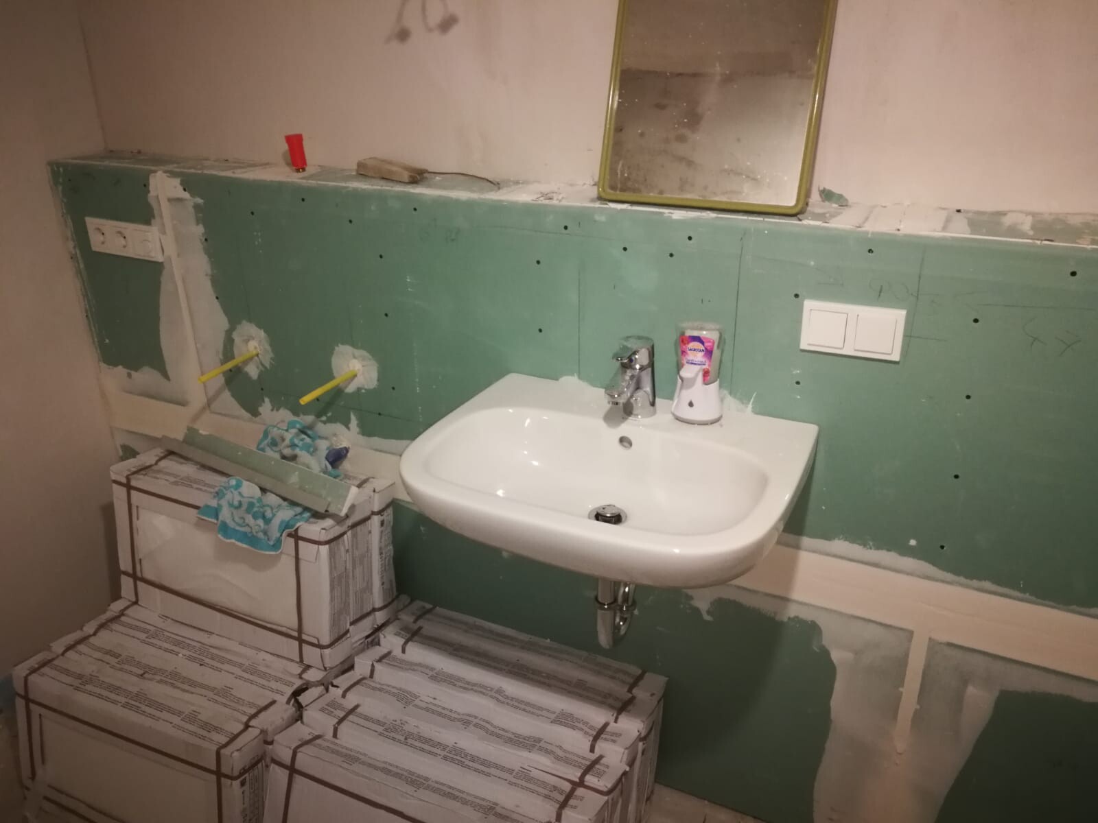 Under renovation: White washbasin on green wall, yellow-framed mirror, construction packages on the floor.