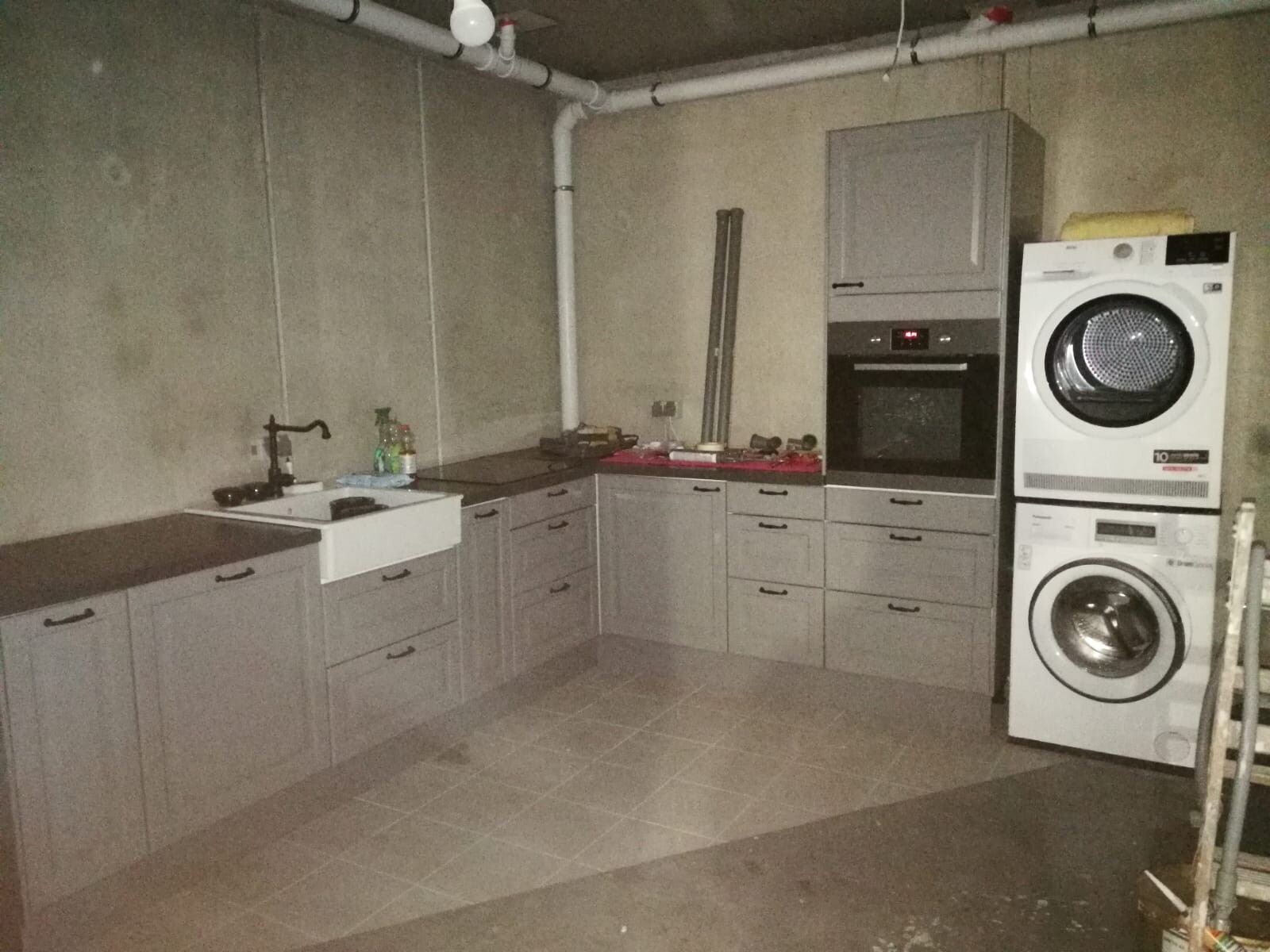 Basement kitchen with gray base cabinets, sink, oven, and stacked washer/dryer.