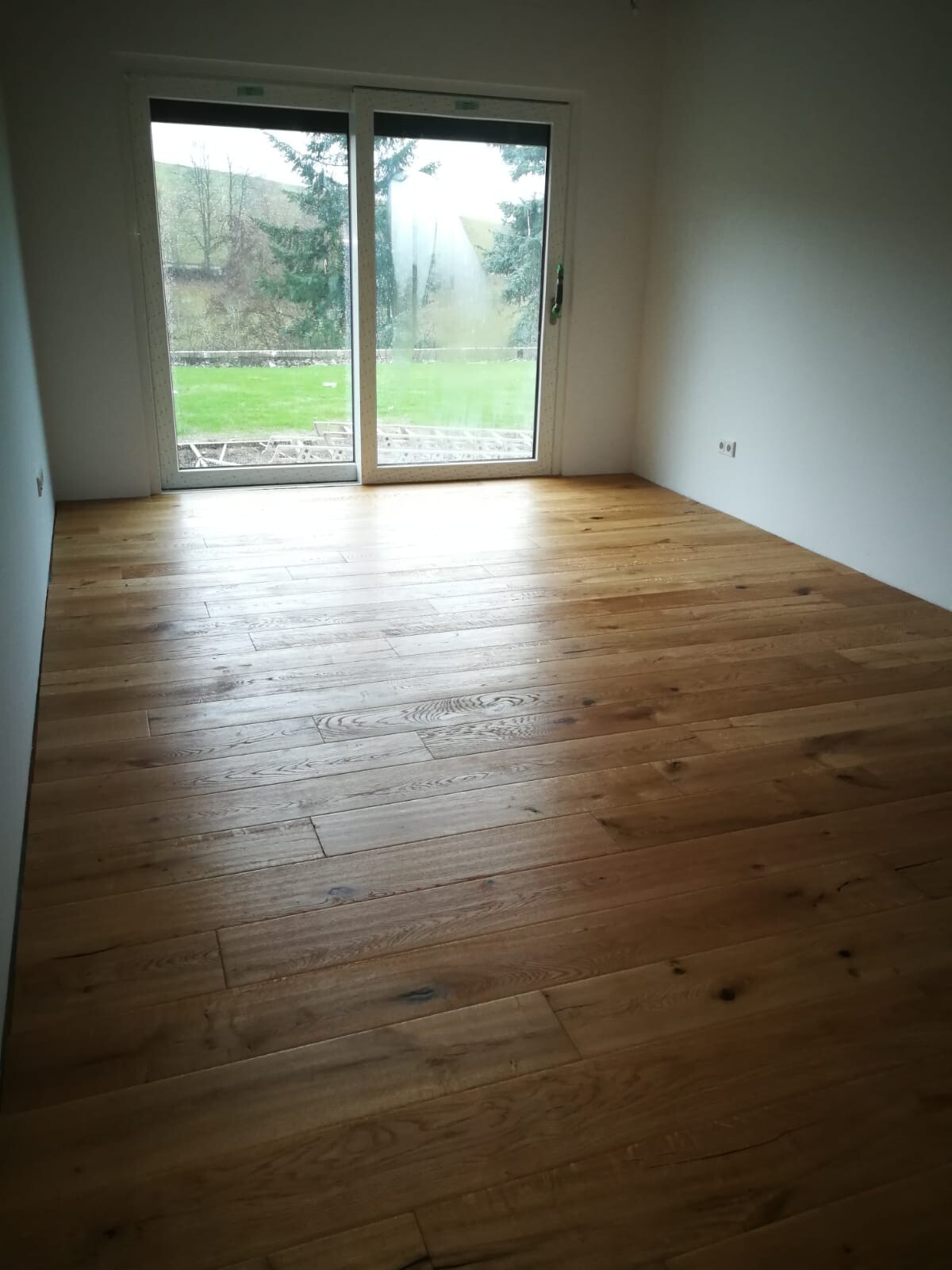 Empty room with wooden floor and large glass doors to the garden, view outside to lawn and trees.