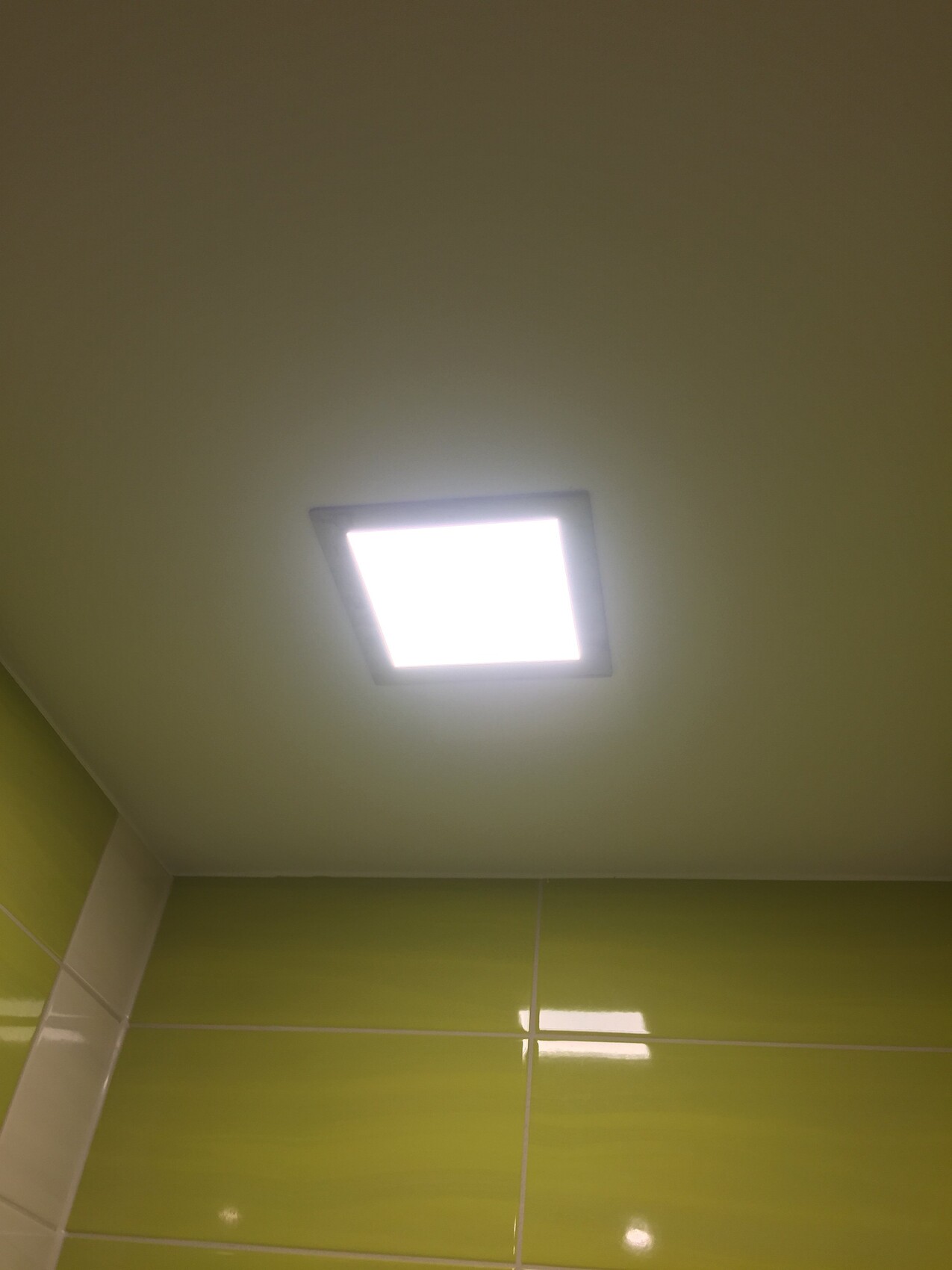 Bright square recessed ceiling light over glossy green bathroom tiles.