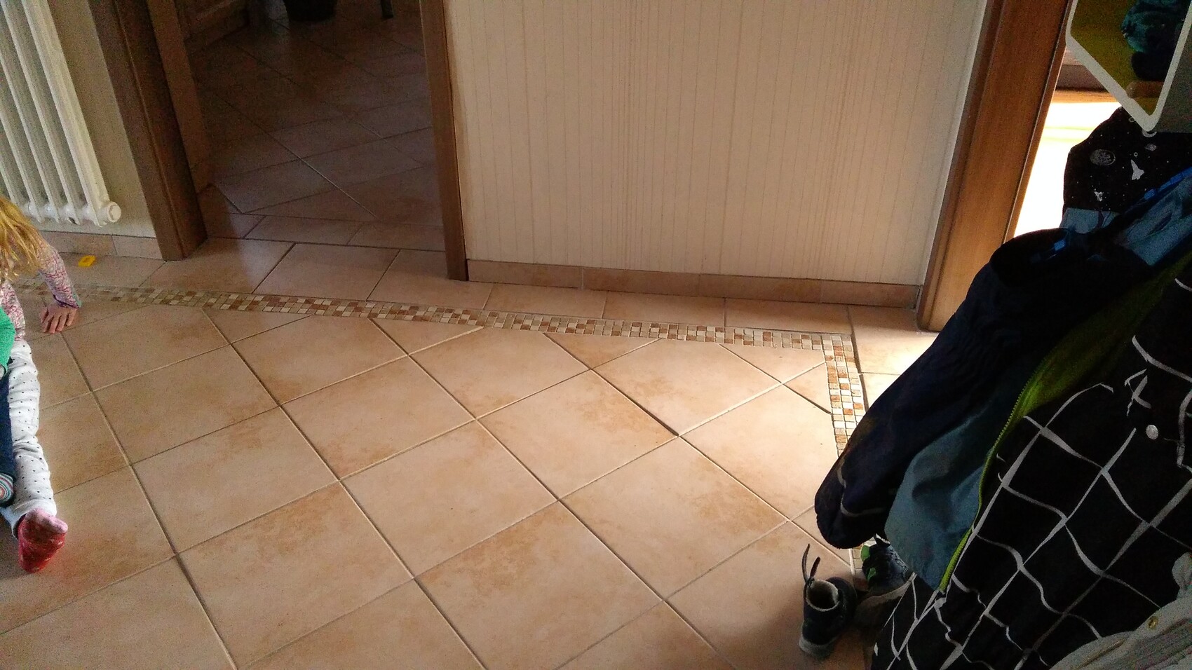Heller Fliesenboden im Flur, linkes Kind halb sichtbar, rechte Garderobe mit Jacken.