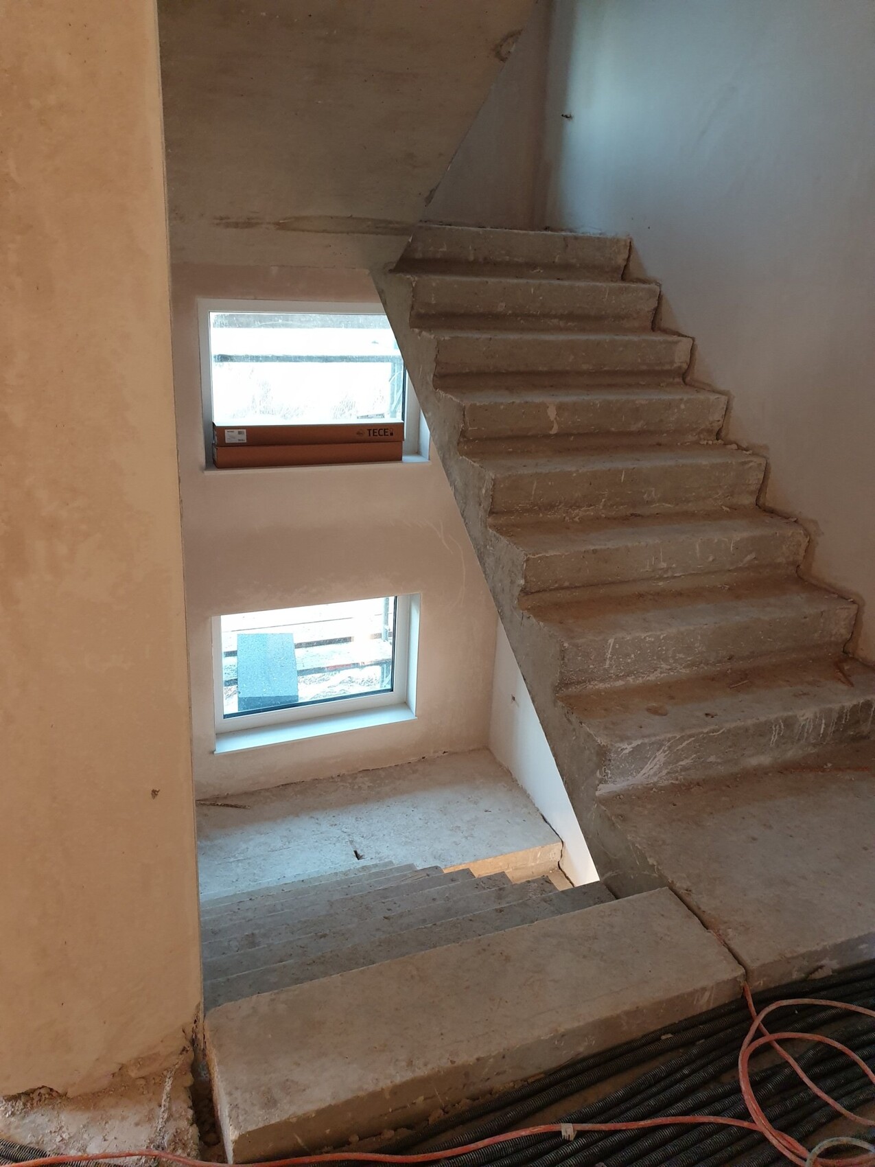 Unbehandelter Rohbau: Beton-Treppe an Innenwand, kleine Fenster, lose Kabel am Boden.