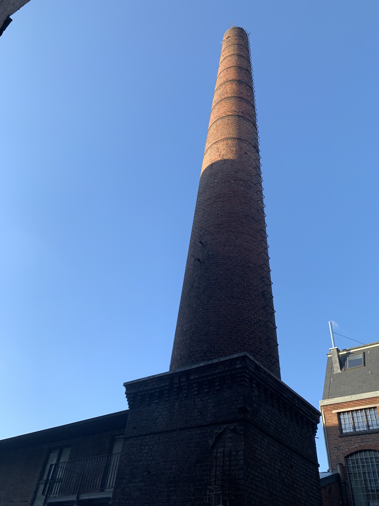 Tall brick chimney next to a factory, external ladder on the side, blue sky.