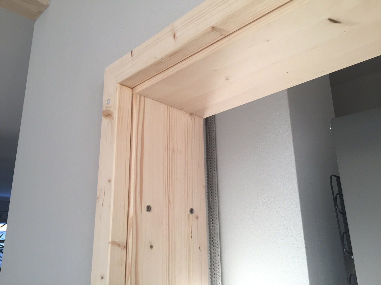 Unbehandelter Holztürrahmen mit Sichtnägeln; graue rechte Wand, Metallführung an Wand.