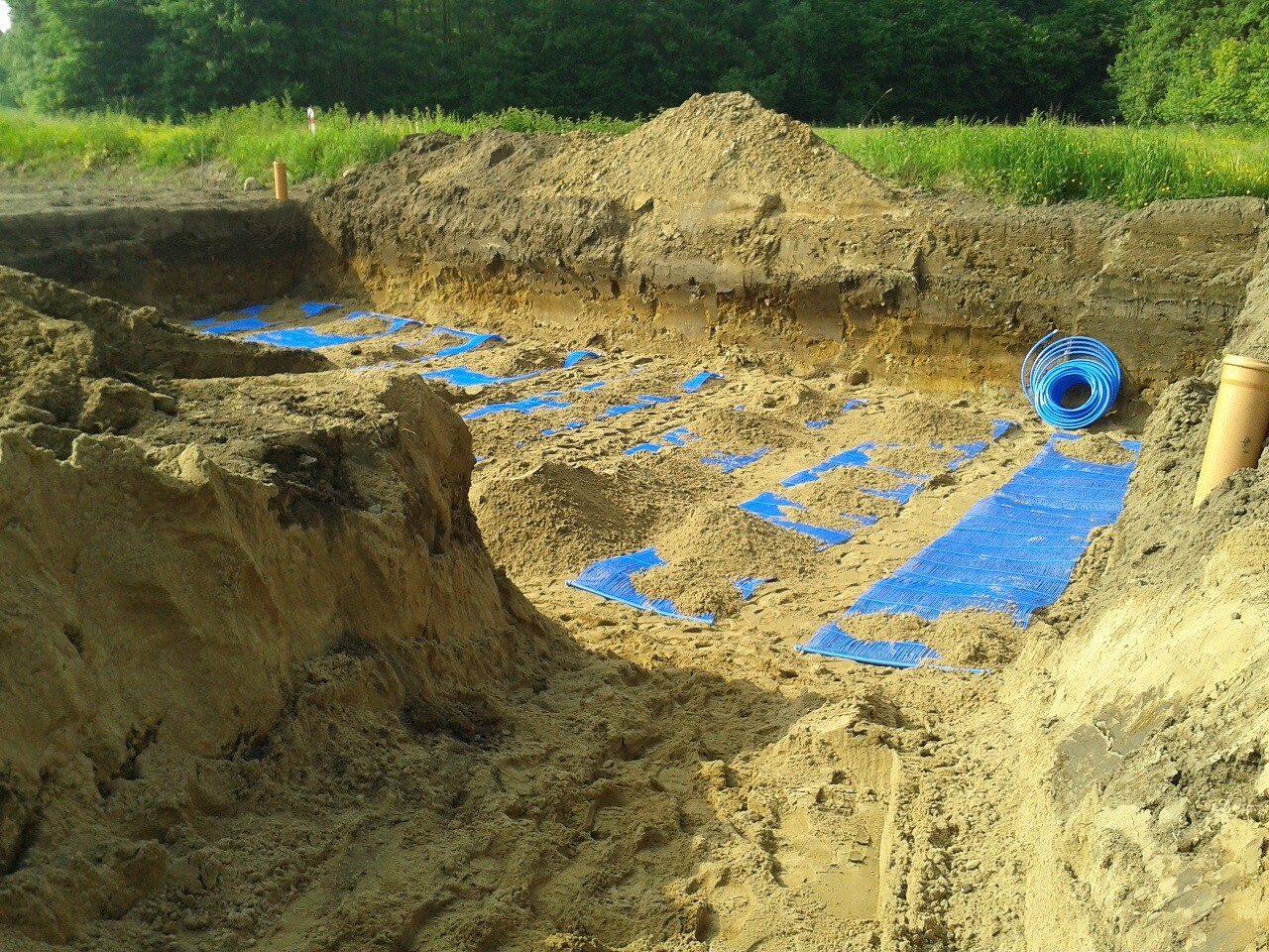 Excavation pit with blue protective mats, piping, and soil at the edge of a green meadow