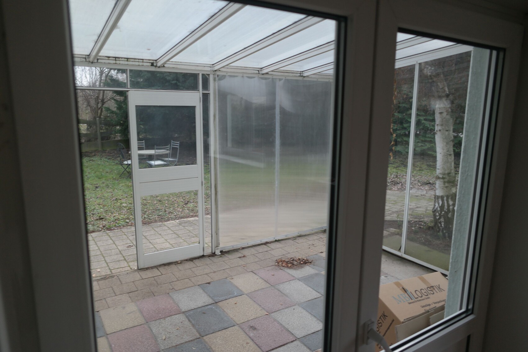 View through window into glazed veranda overlooking garden with furniture; cardboard on floor.