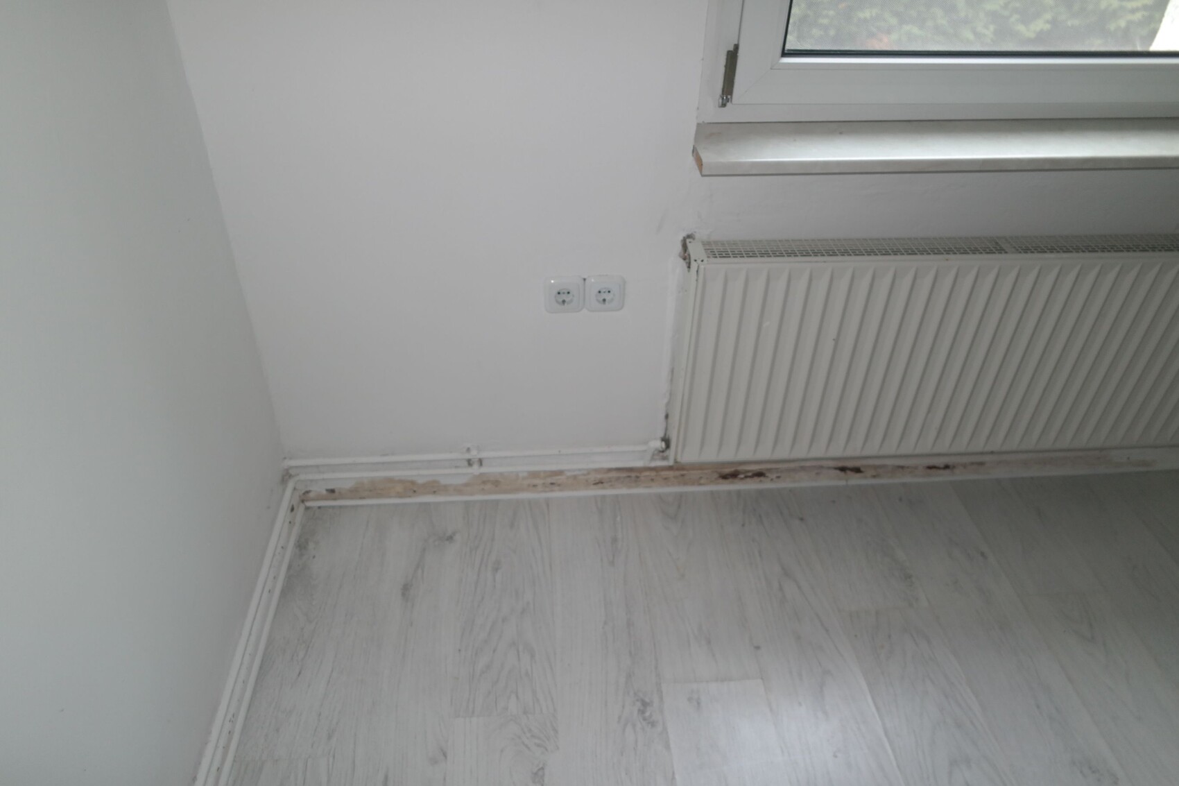 White room corner with window above radiator; two electrical outlets; damaged baseboard.