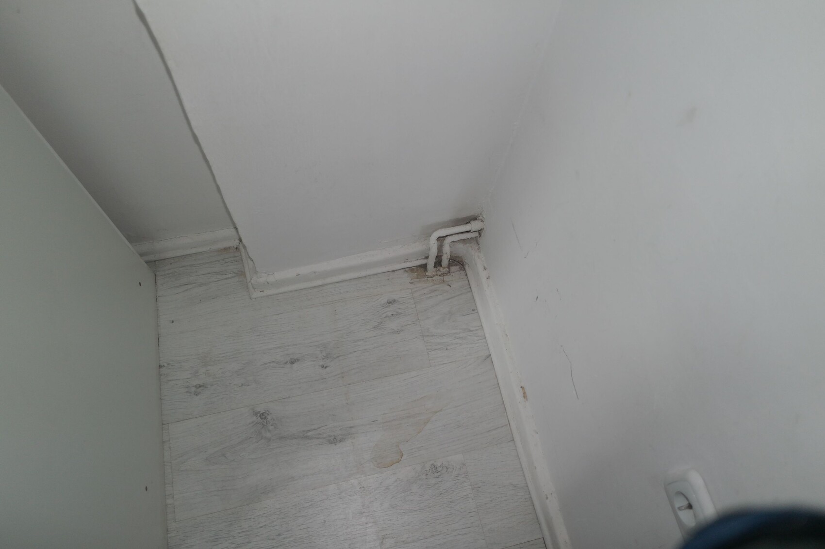 Corner view of a room: white walls, gray laminate floor, short pipe on floor.