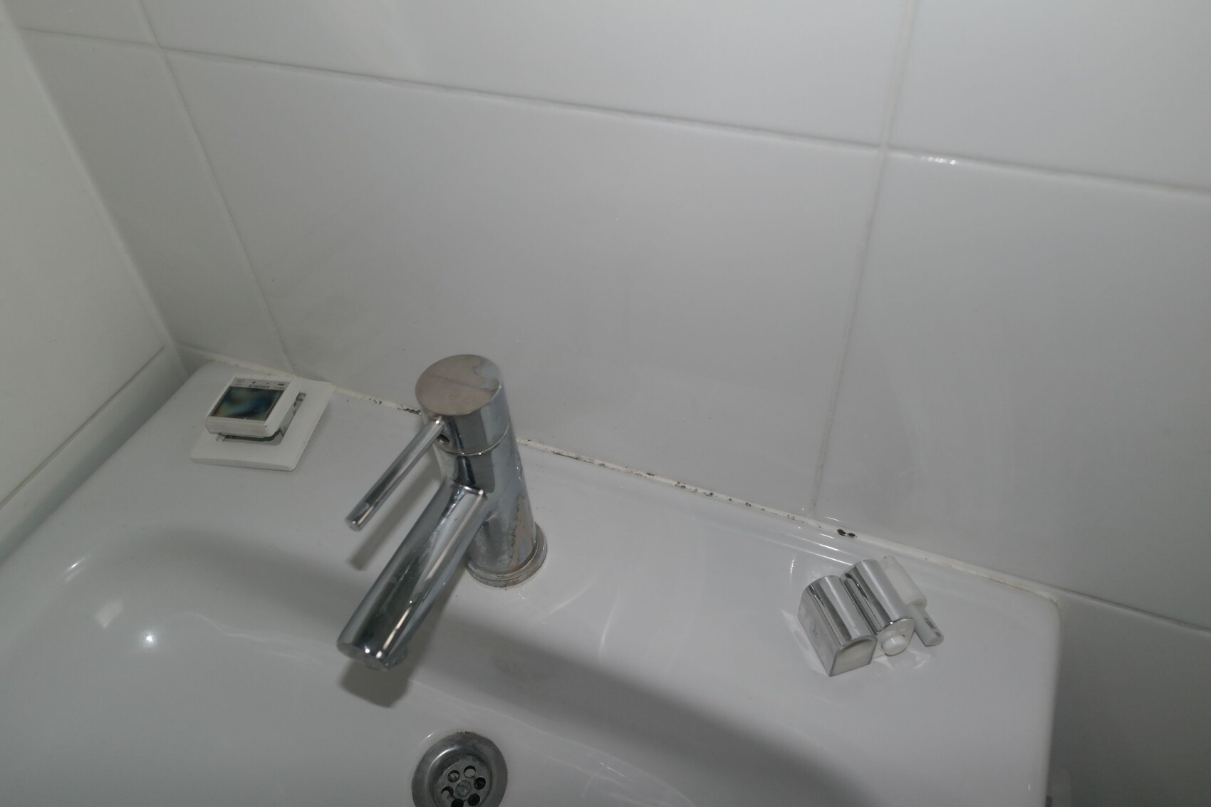Sink with chrome faucet; to the left, a small digital clock on the shelf; white tiles in the background.
