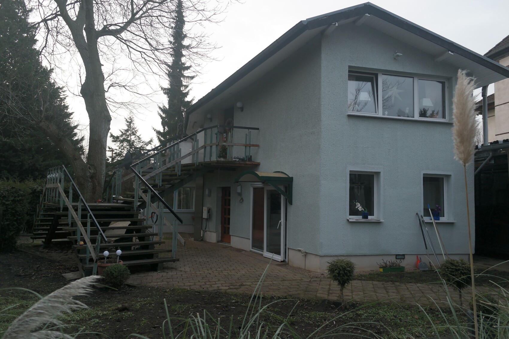 Two-story light blue house with exterior stairs and balcony, garden in front.