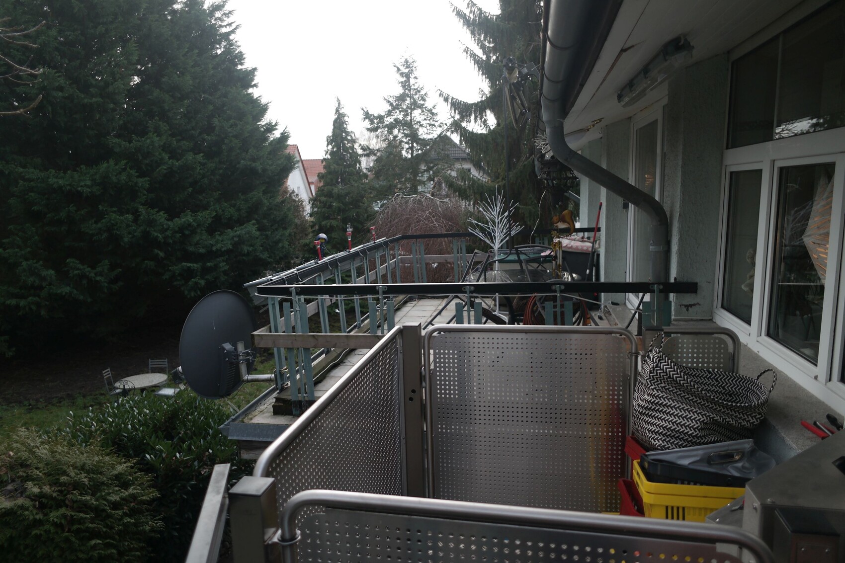 Balcony with perforated metal railing, satellite dish, boxes and garden tools in front of houses.