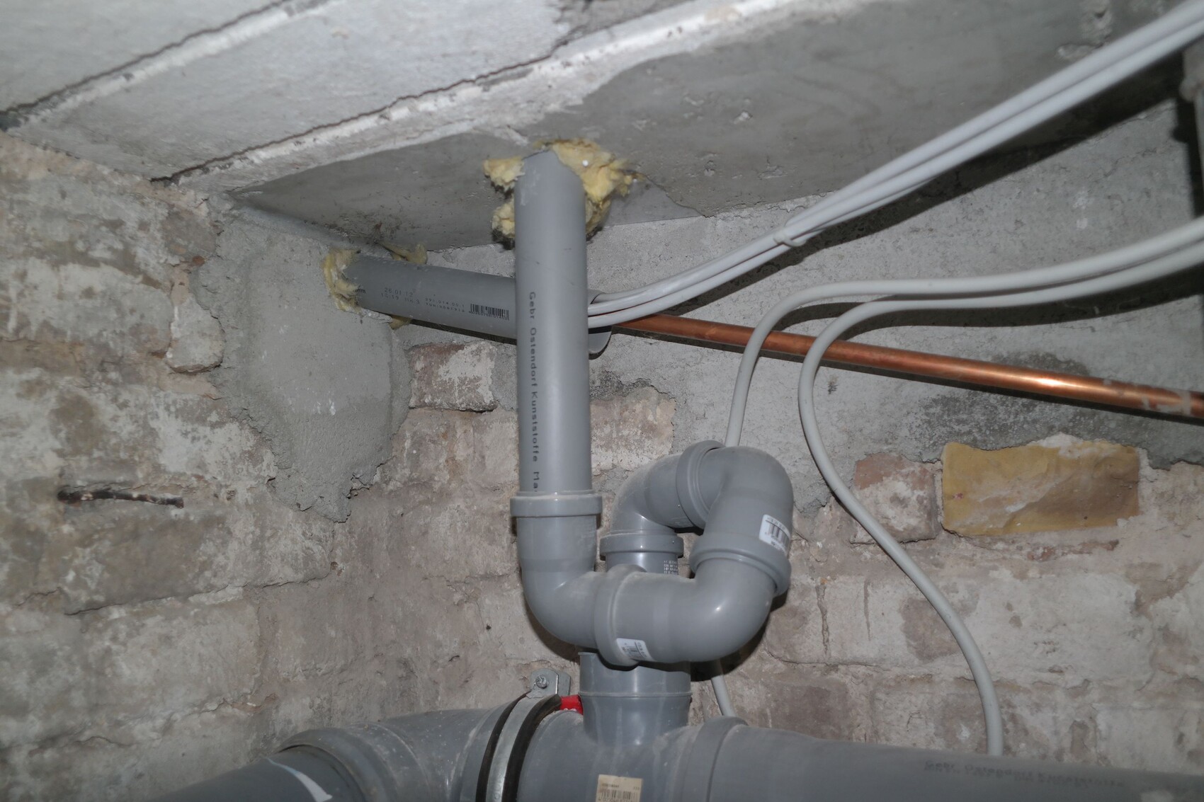 Basement room with gray PVC drain pipes, siphon, copper pipe, and cables on rough stone wall.