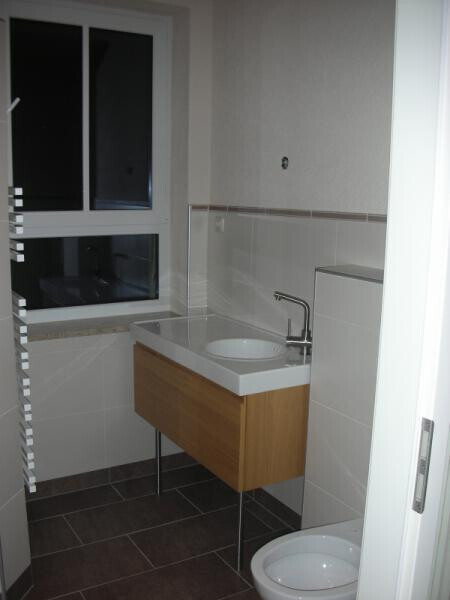Small bathroom with washbasin on wooden cabinet, toilet, window, and tiled floor.