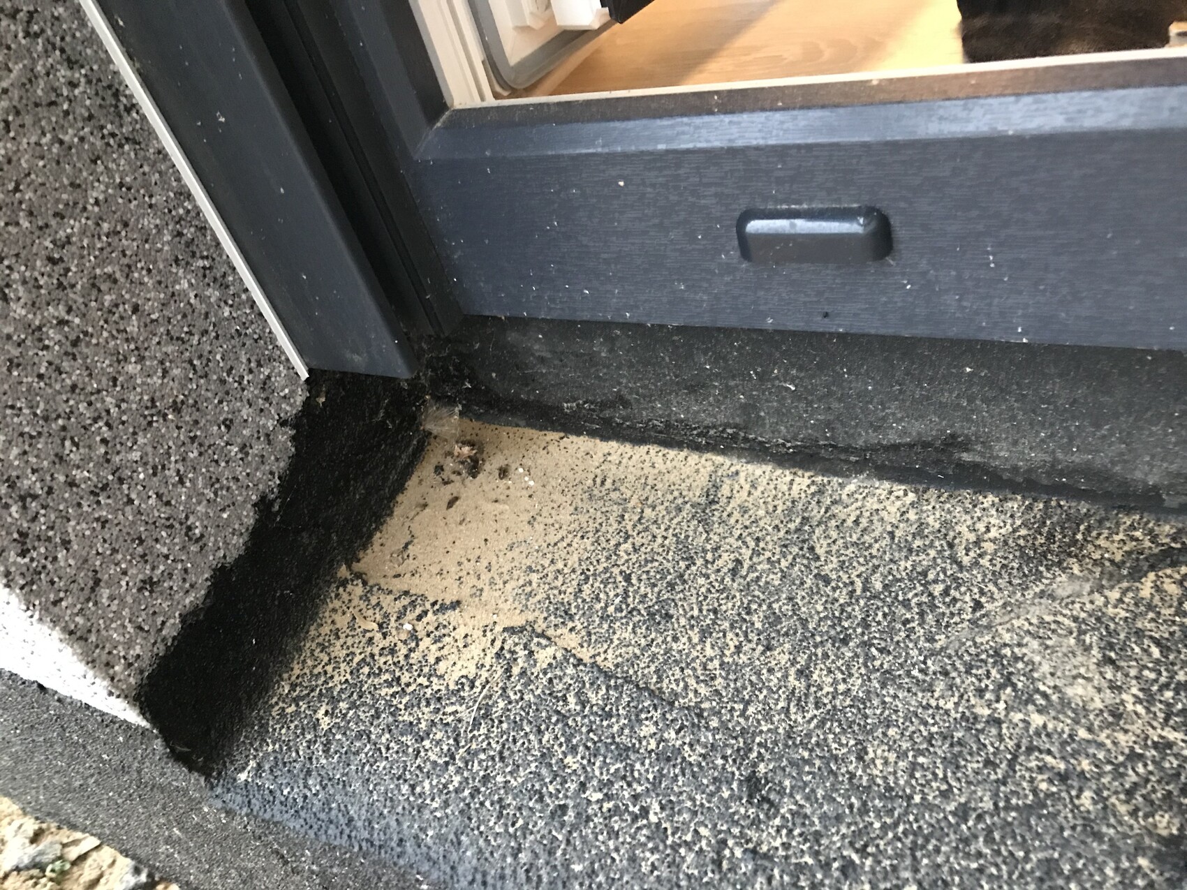 Exterior area of a door threshold with sand on a dark slab; wooden floor visible inside.