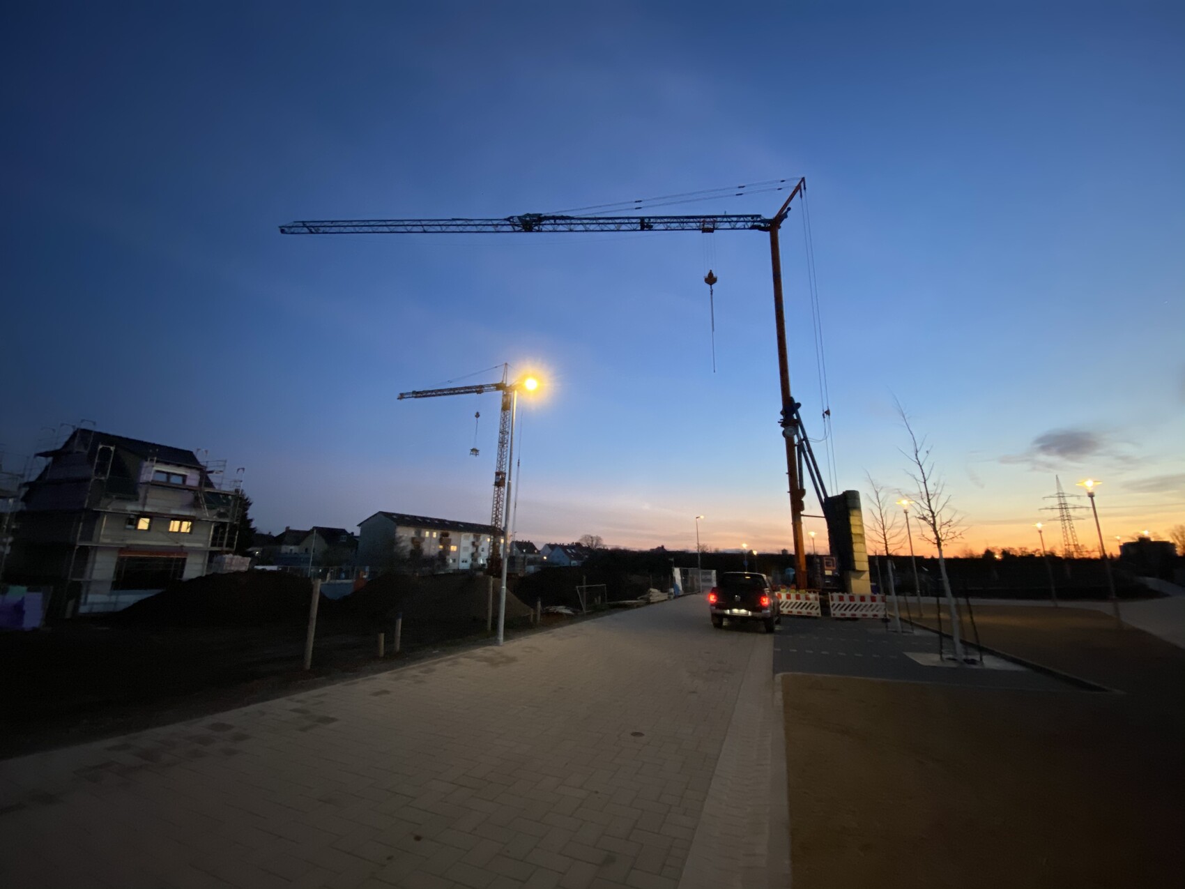 Evening construction site photo: cranes, vehicle, construction fences, street lamps, skyline at dusk.