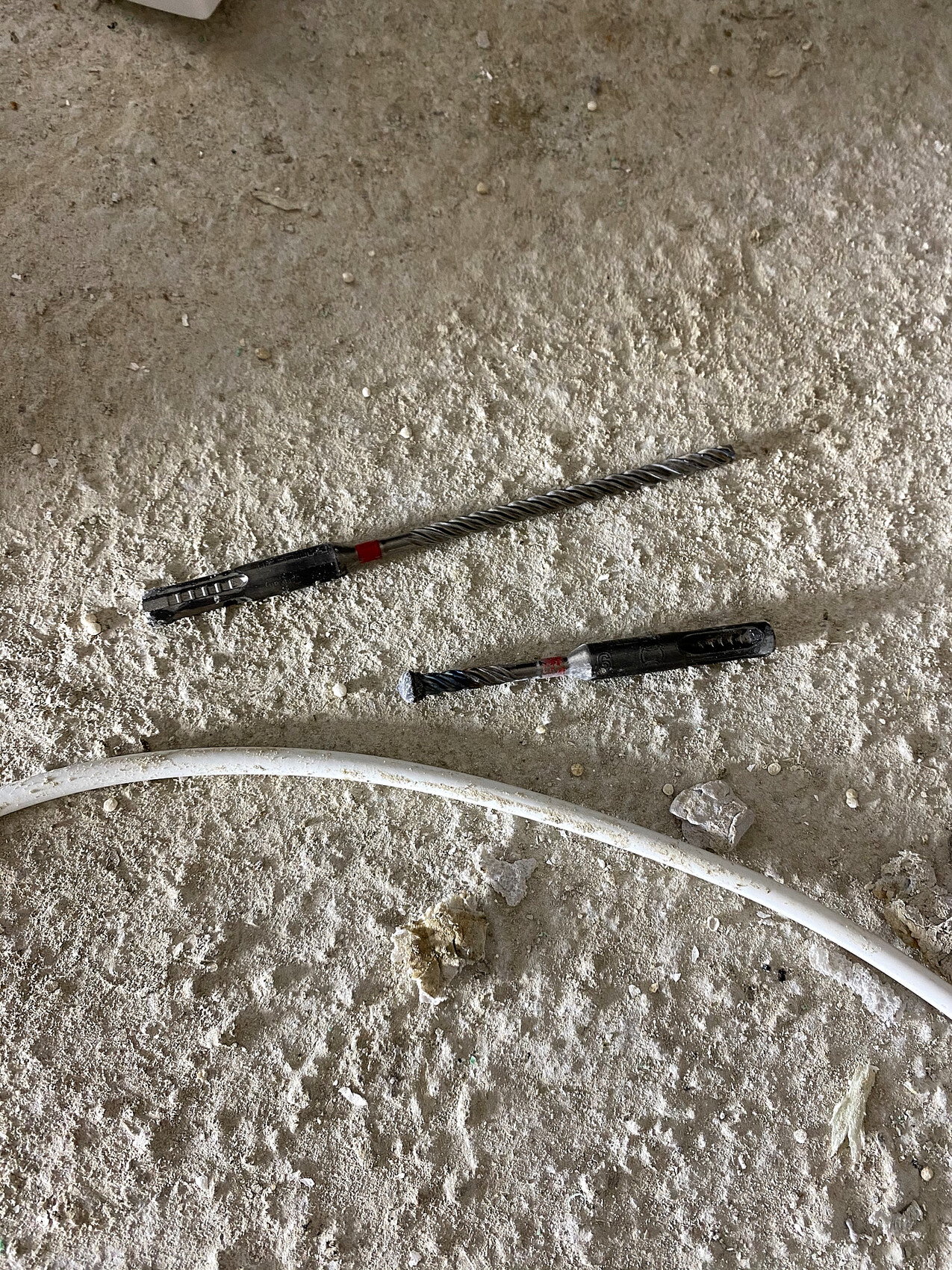 Two spiral drill bits lying on dusty concrete floor next to a white pipe.
