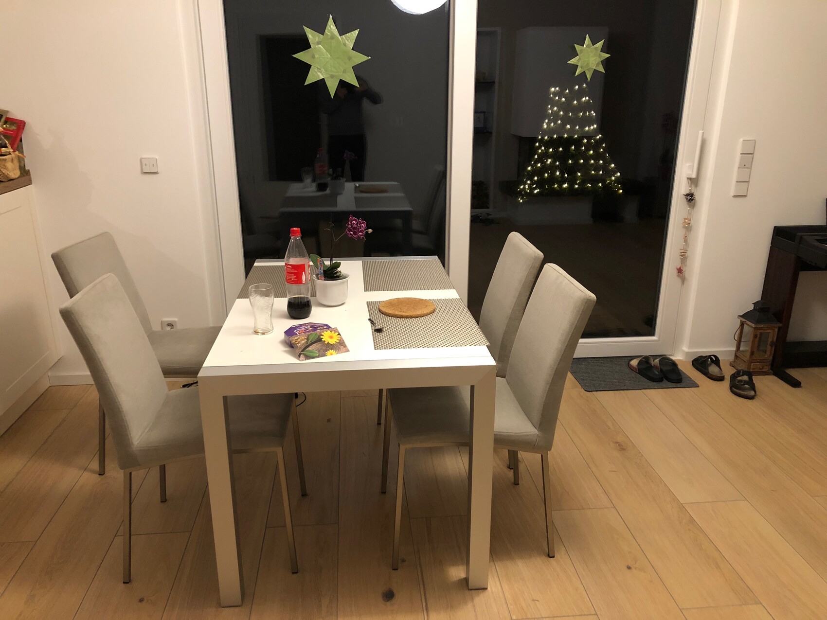 White dining table with four chairs, soda bottle, plant; window with stars and Christmas tree.