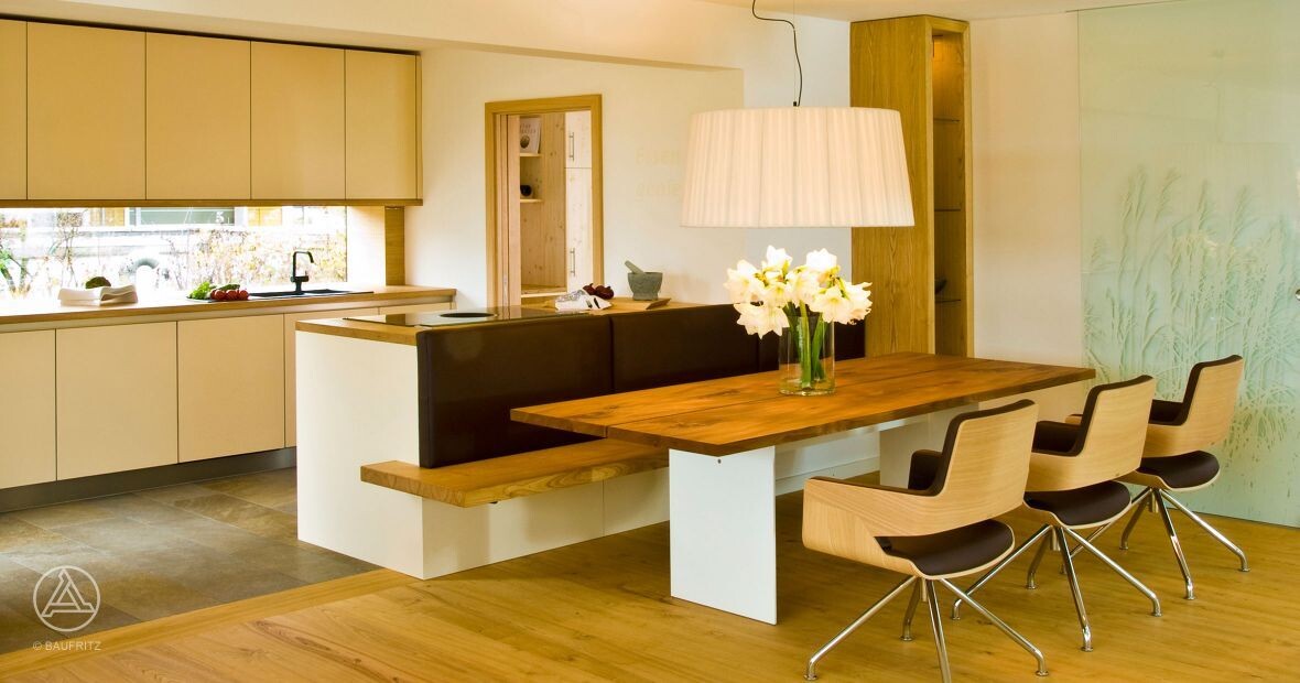 Modern kitchen-dining area: long wooden table with bench and chairs, vase with flowers, bright interior.