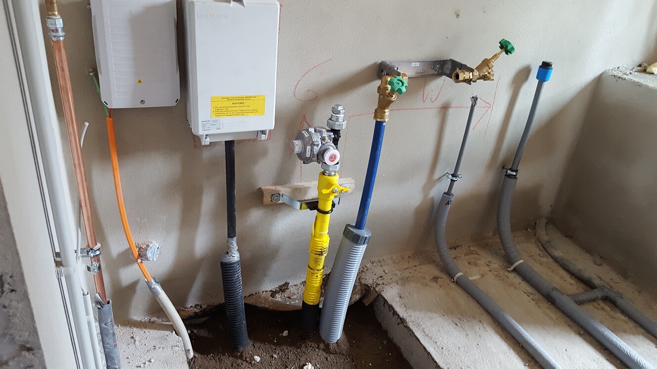 Gas- und Wasserleitungen mit Ventil, Messgerät und flexiblen Schläuchen an Wand.