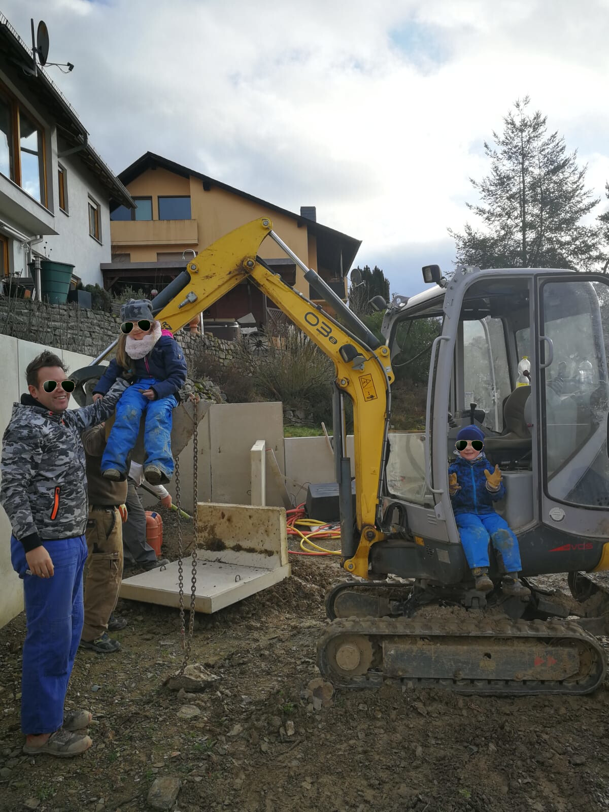 Familie auf Baustelle: Gelber Minibagger, ein Kind im Fahrerhaus, ein Kind wird hochgehoben.