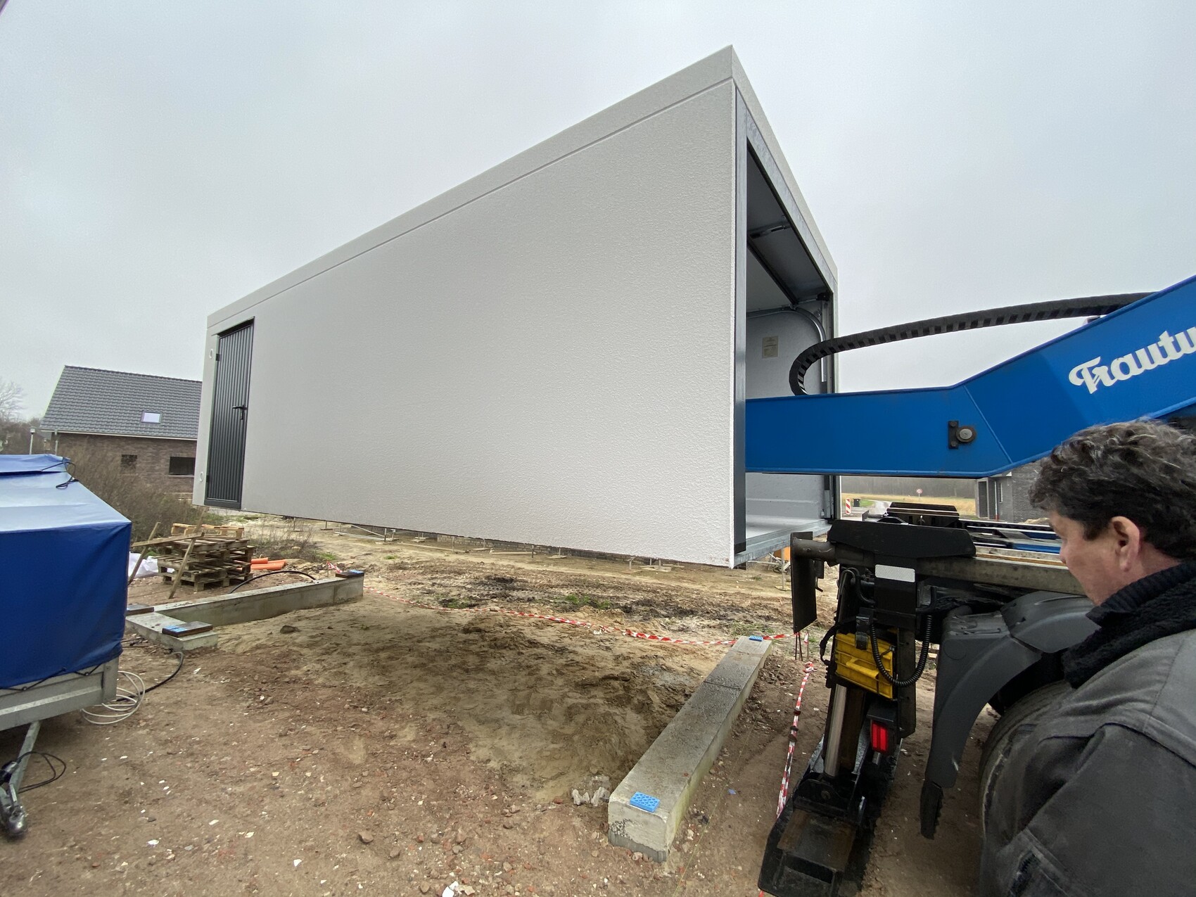 White modular construction container being lifted onto a foundation by a blue crane; construction worker on the right.
