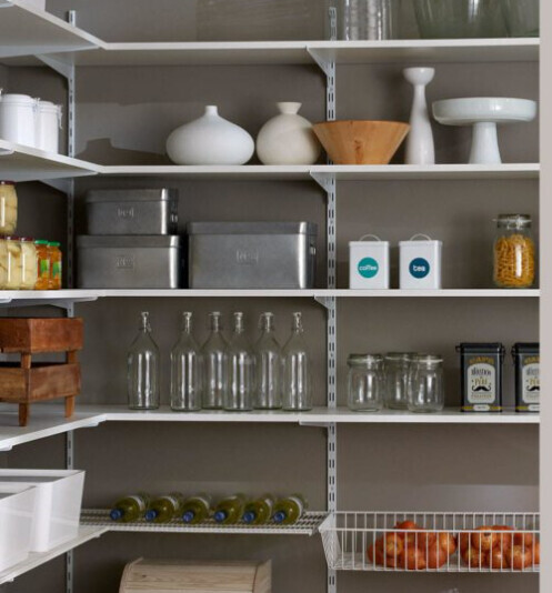 Open kitchen shelf with glasses, metal boxes, coffee and tea canisters, fruit basket with oranges.