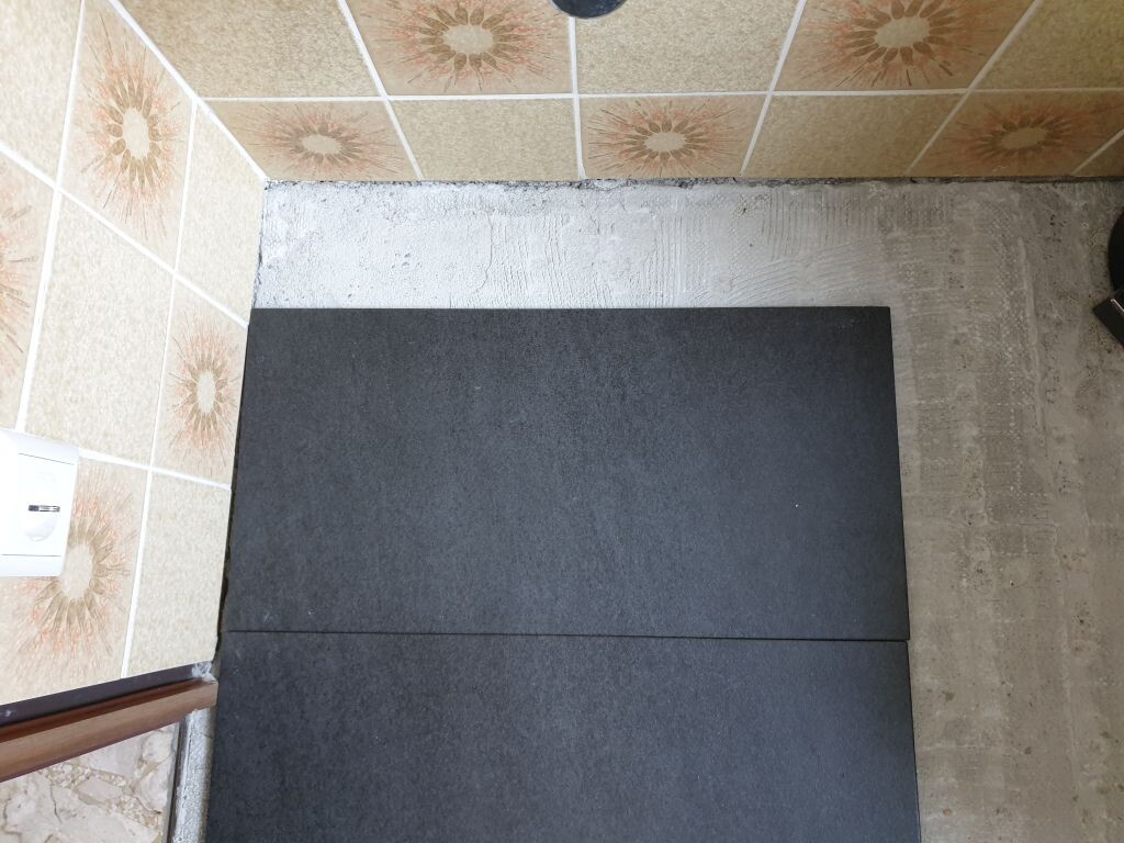 Two dark gray tiles lying on a concrete floor next to patterned wall tiles; socket visible.