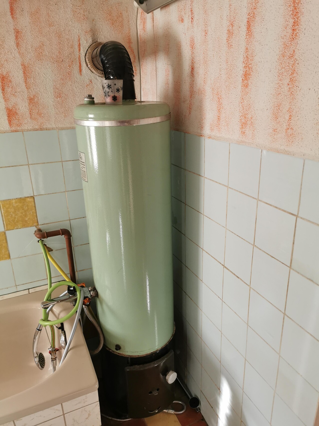 Grüner Wasserboiler in Badezimmerecke mit Rohren, Duschschlauch und orange Wandflecken.
