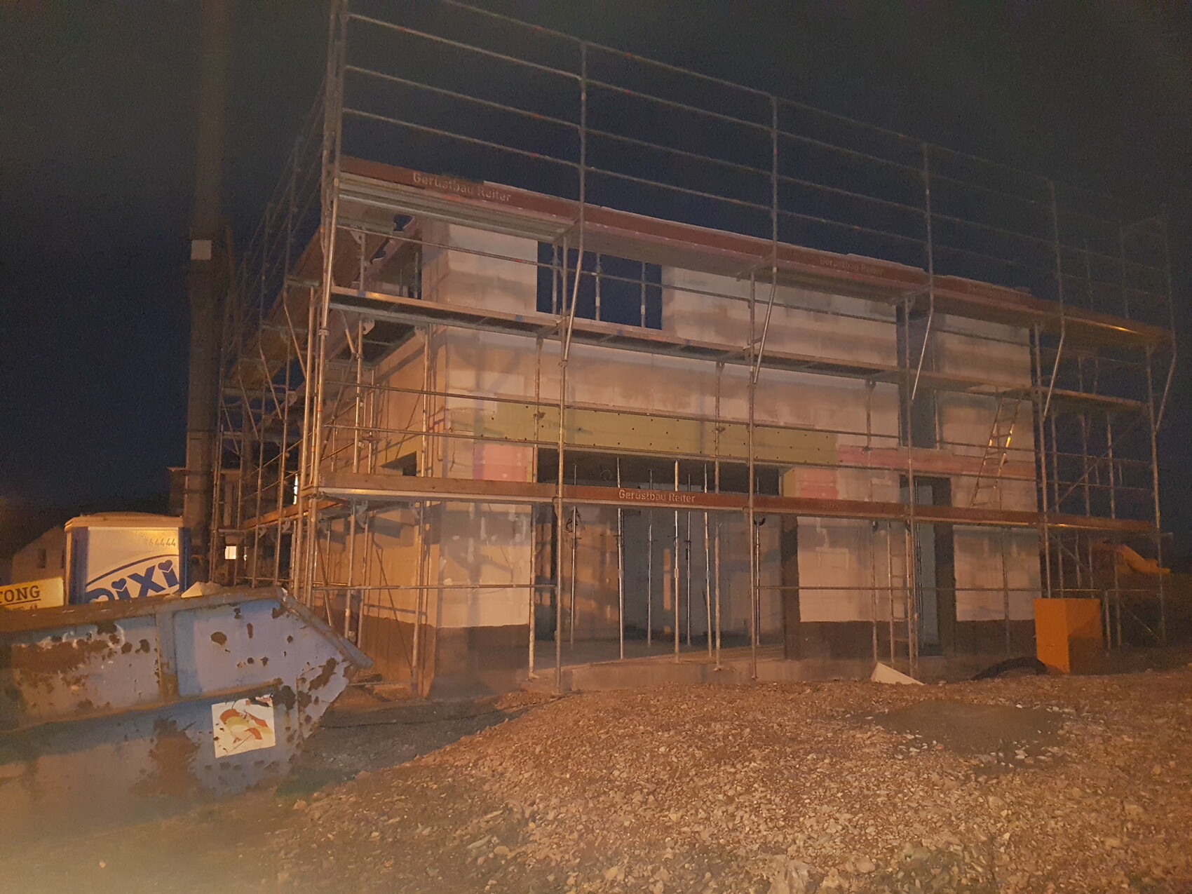 Construction site at night: multi-story scaffolding around a building, gravel ground in front, waste container on the left.