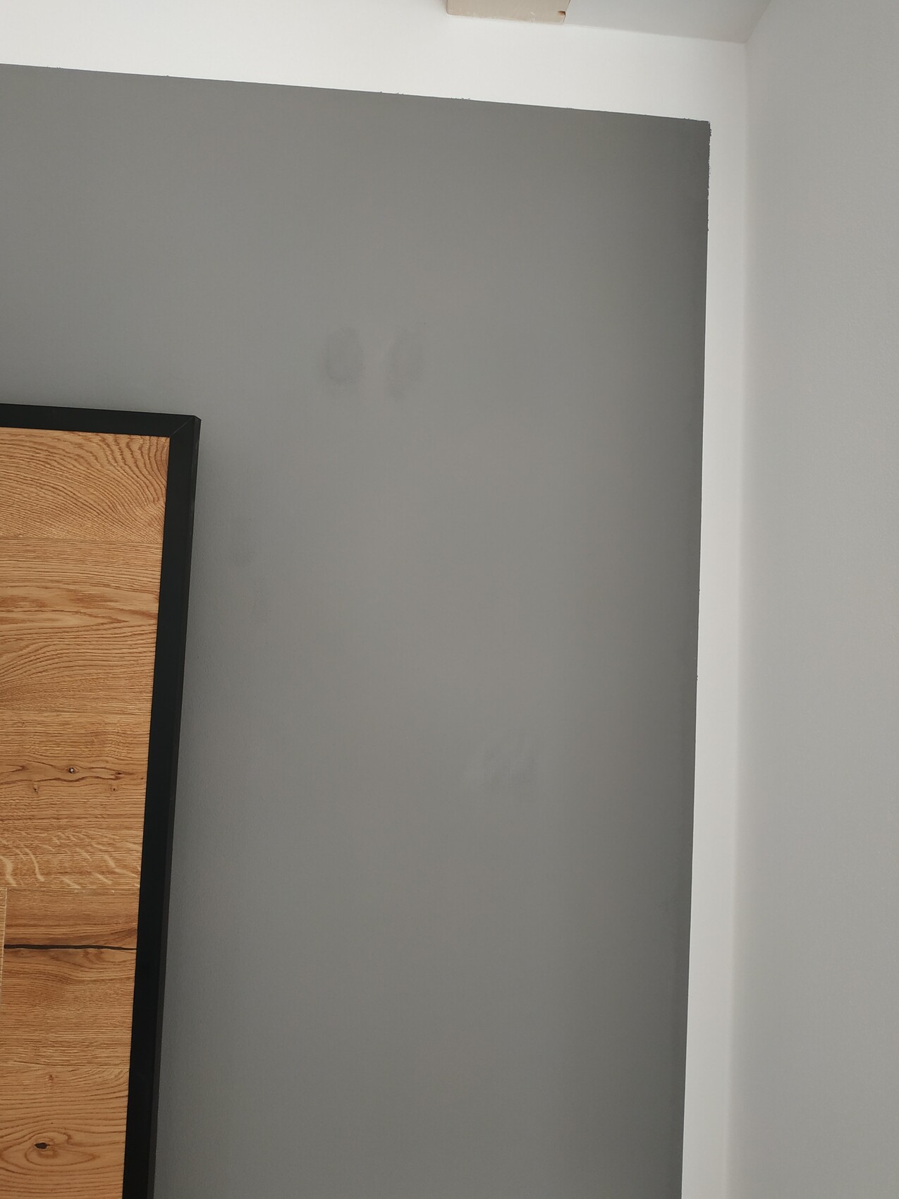 Graue Innenwand mit links sichtbarem Holzpaneel und schwarzem Rahmen; weiße Decke.