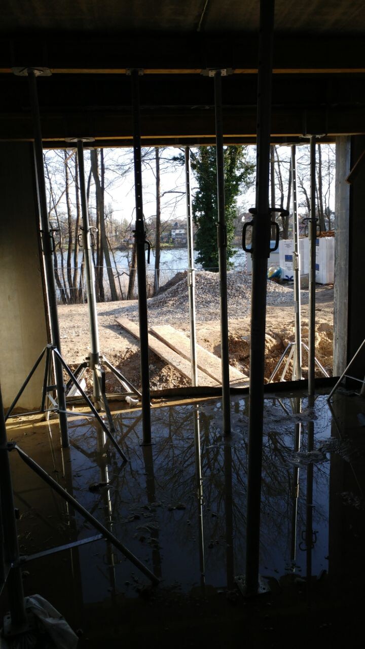 Interior view of a construction site with supports, a puddle of water, and a view outside to trees by the water.