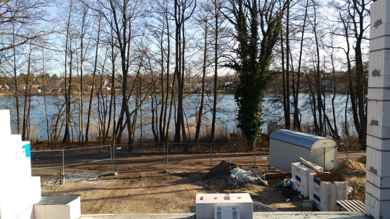 Construction site at the riverbank: fence, construction container, pallets, and trees in the background.