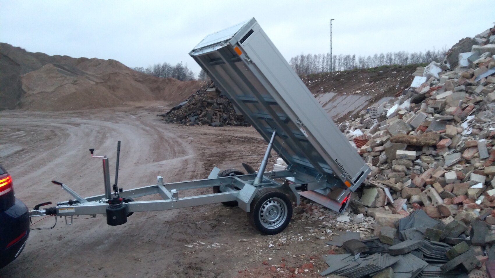 Single-axle tipper trailer attached to a car, tipping the bed, construction debris visible.