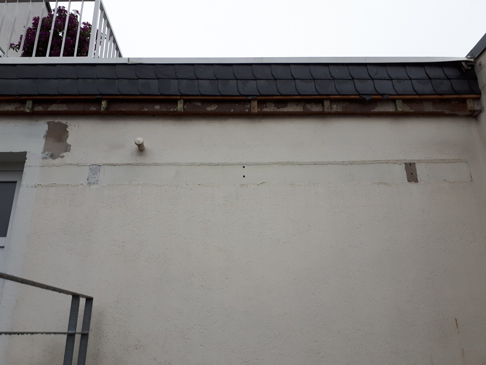 Weiße Außenwand mit rauer Putzstruktur, dunkles Schieferdach, Balkon mit violetten Blumen