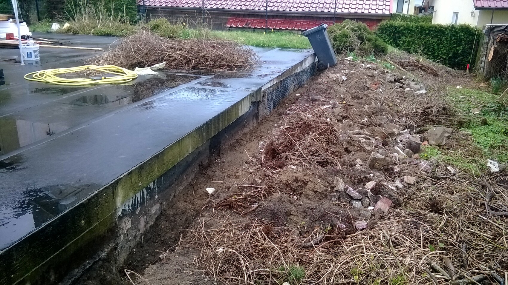 Wet concrete slab with yellow hose, branches and debris next to a waste container.