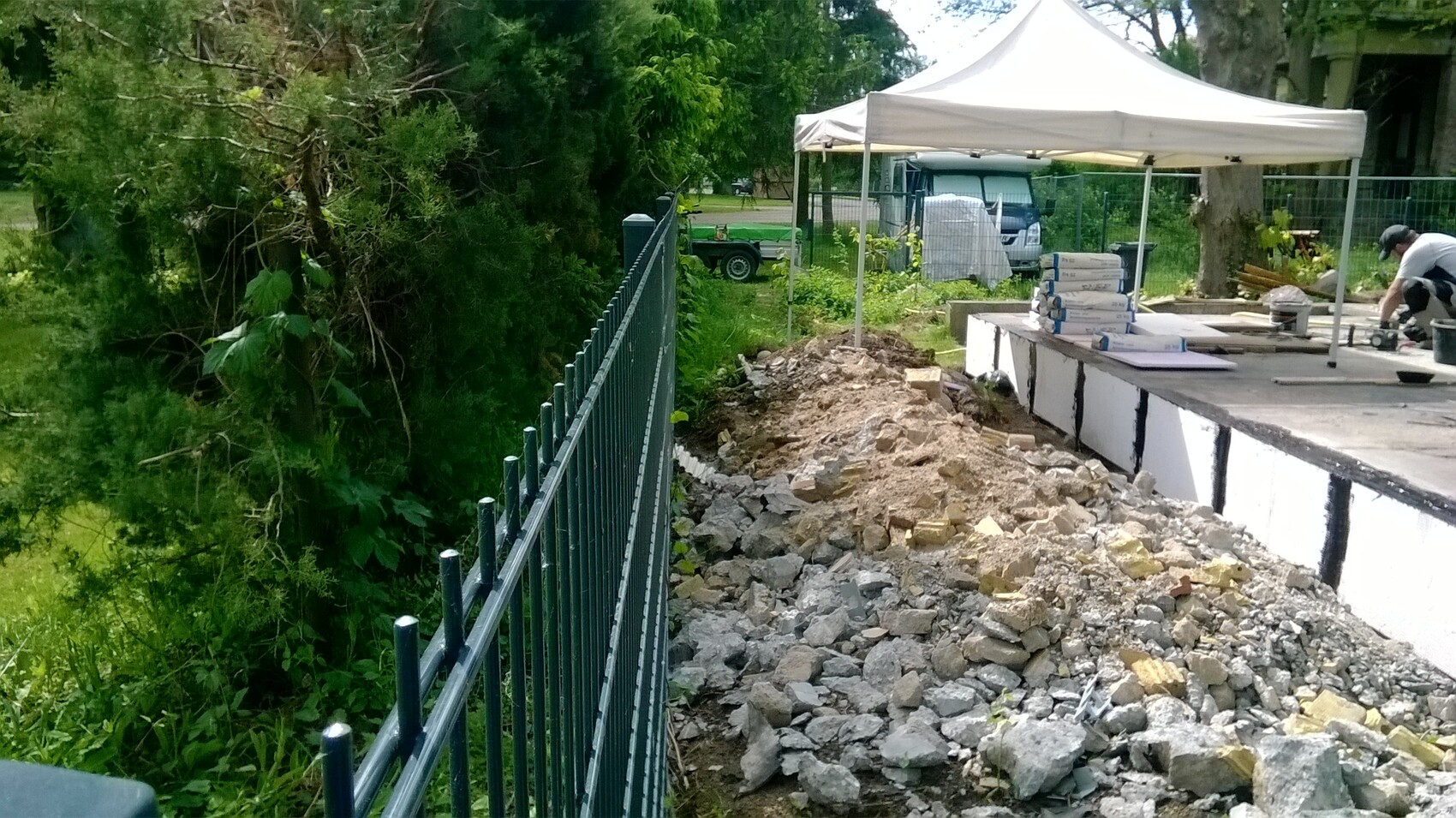 Baustelle: Geländer links, Haufen Kies und Geröll, Pavillon, Arbeiter mit Baumaterial.