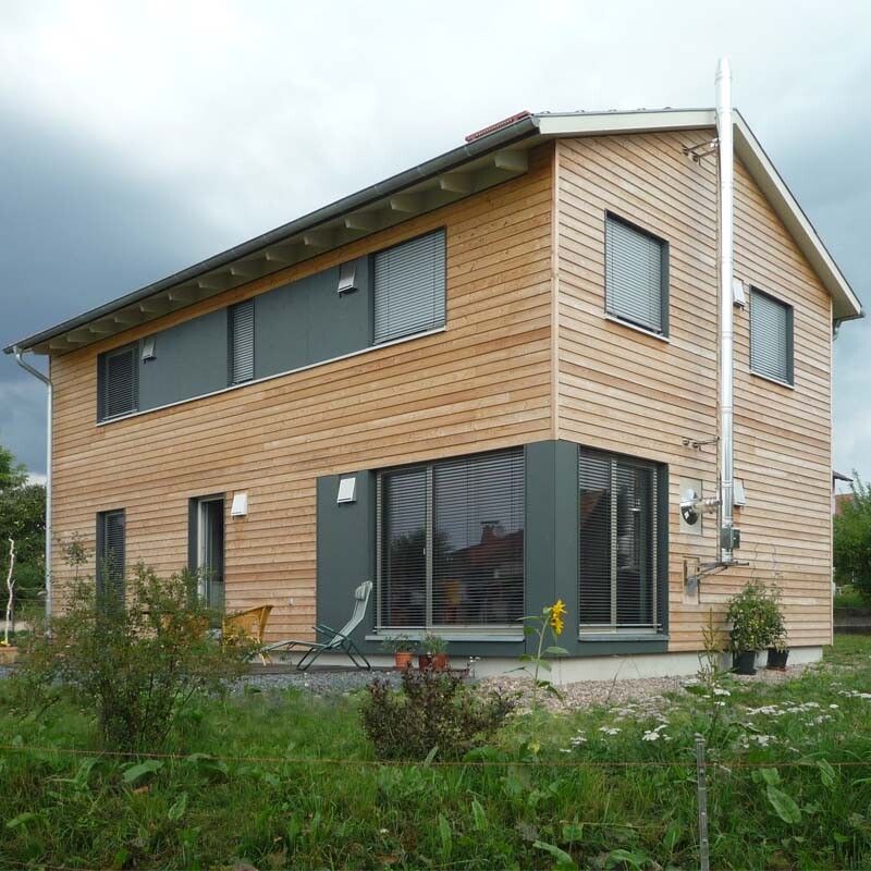 Two-story wooden house with large windows, wood cladding, and garden.