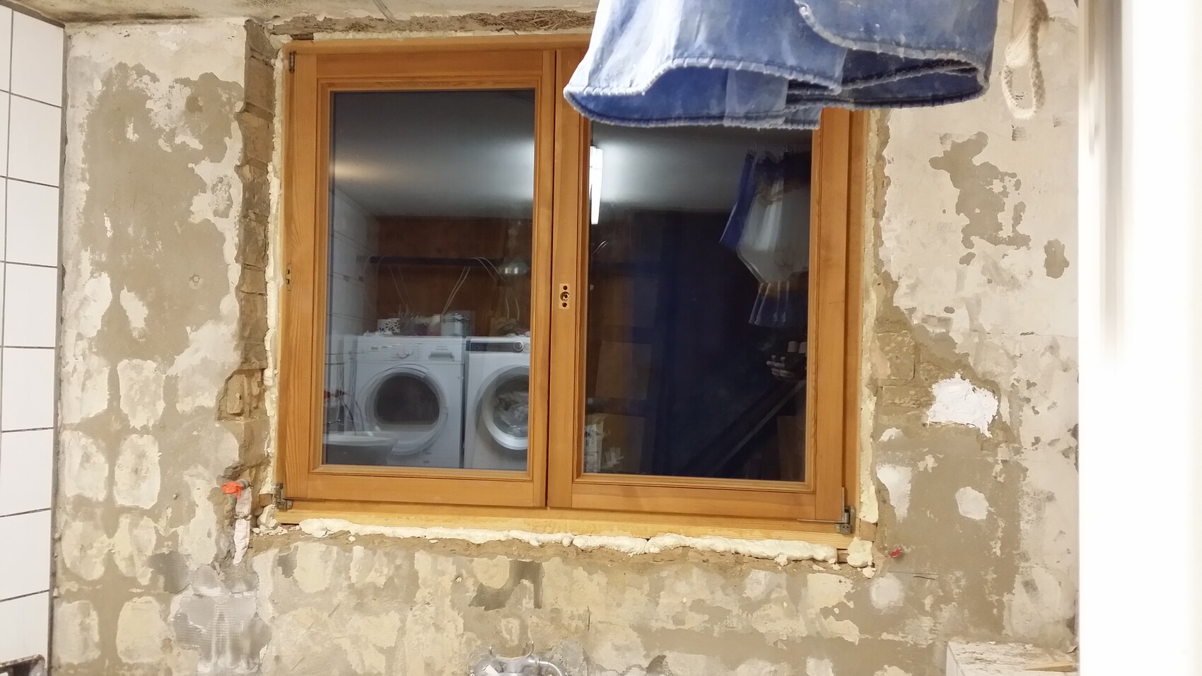 Wooden window in renovated, rough wall; behind the glass, two front-loading washing machines are visible.