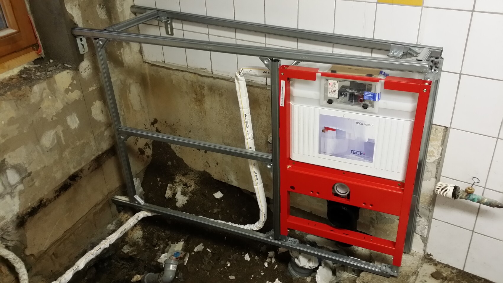 Wall-hung toilet frame made of metal with red flushing cistern in front of tiled bathroom wall.