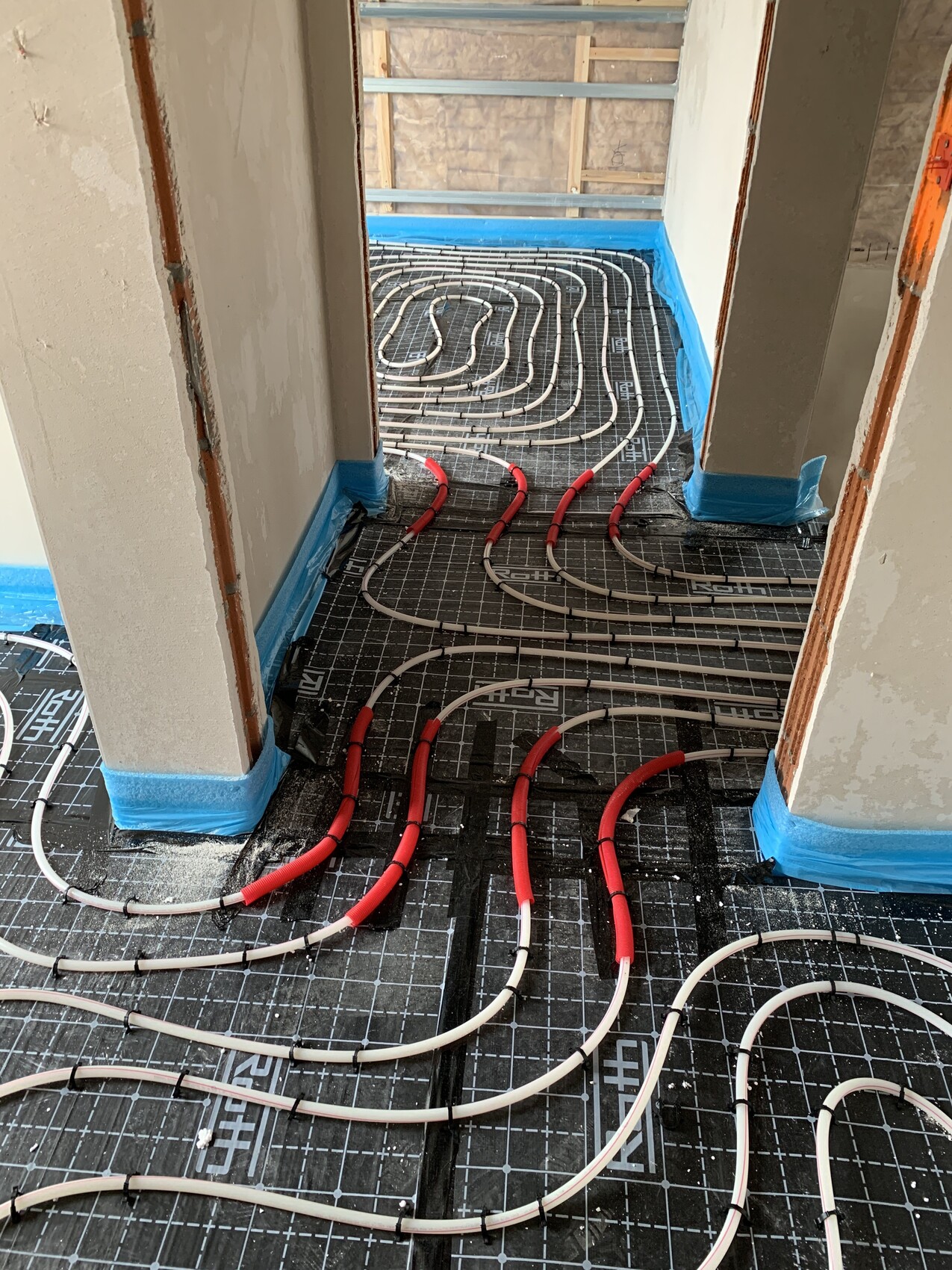 Underfloor heating: red and white pipe network laid on black grid panel in construction site.