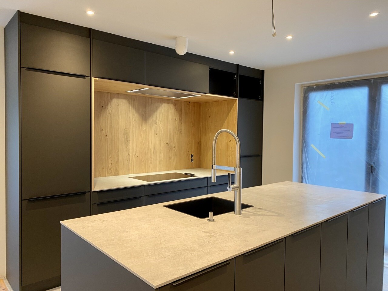 Modern kitchen with black fronts, wooden backsplash behind the cooktop, island with sink and faucet.