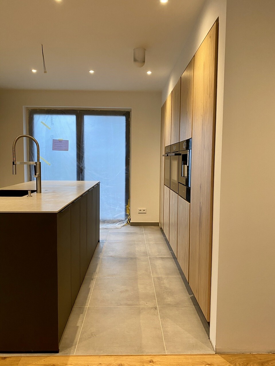 Modern kitchen with island on the left, sink/faucet; wooden cabinets with ovens on the right, window wrapped.