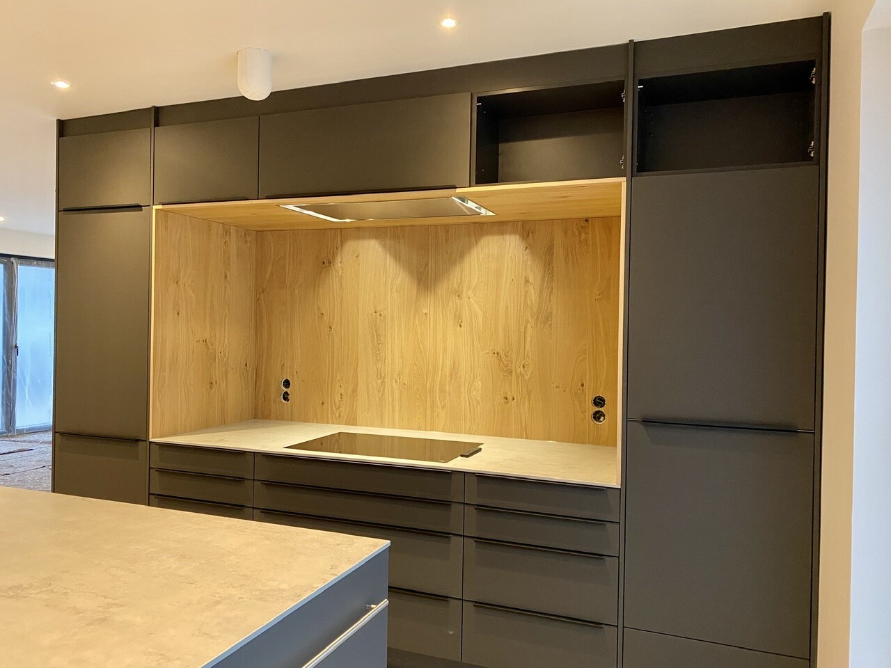 Modern kitchen line with dark gray fronts, wooden wall, induction cooktop and many drawers.