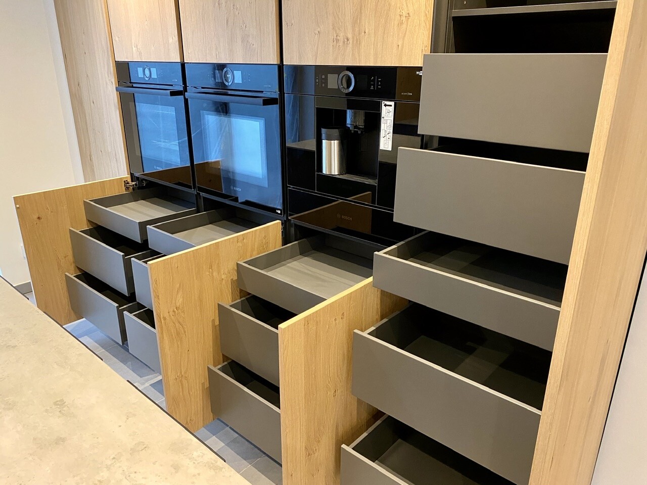 Modern kitchen with integrated Bosch ovens and many open wooden and gray drawers.