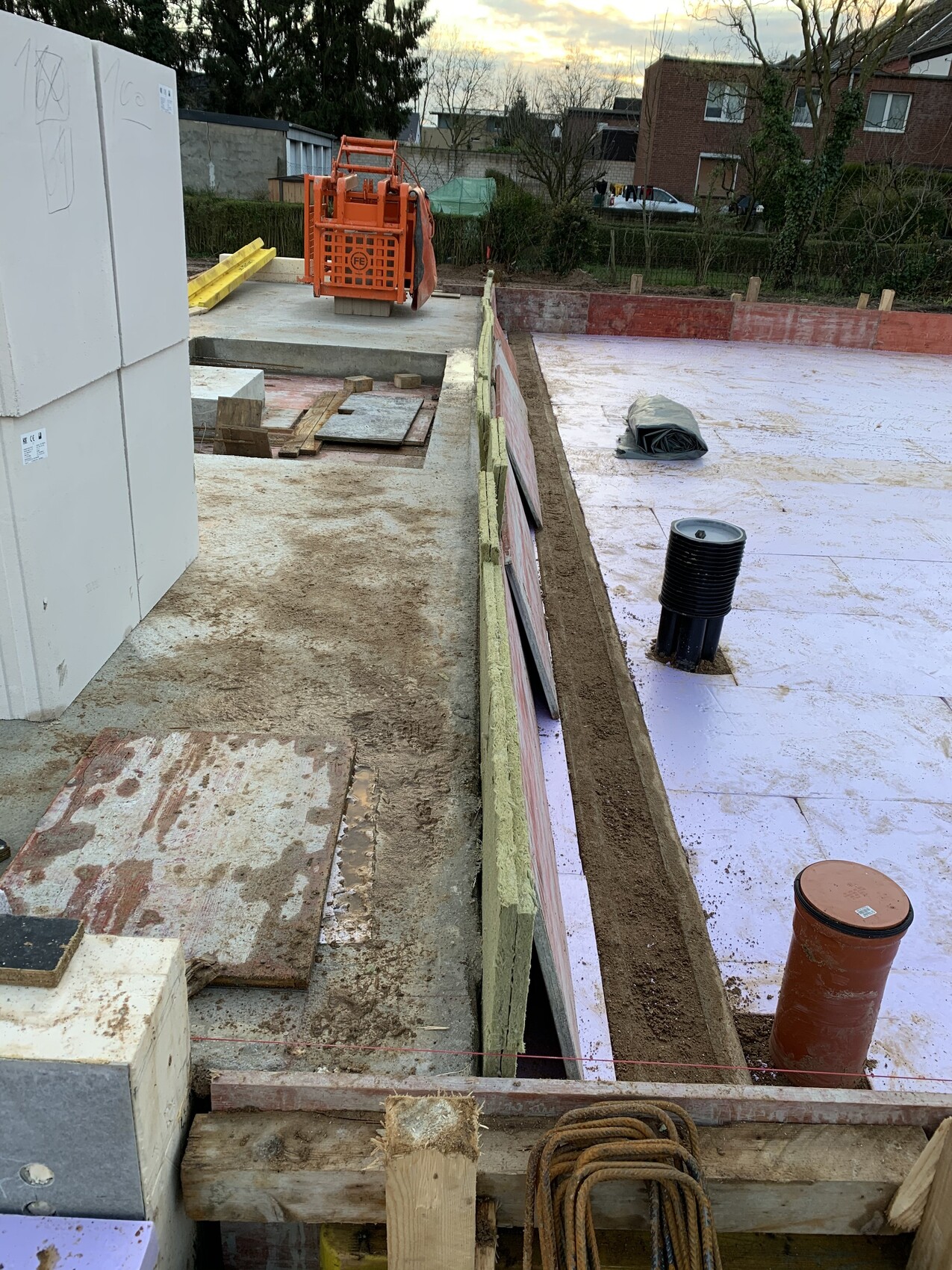 Construction site: foundation trench with pink waterproofing membrane, orange construction machine, wooden boards.